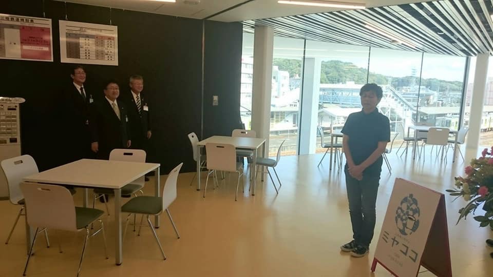 1つのテーブルに4脚の椅子がセットで置かれた施設で、壁際に市長と男性2名が立ち、「ミヤココ」と書かれた看板の横に女性が1名立っている写真