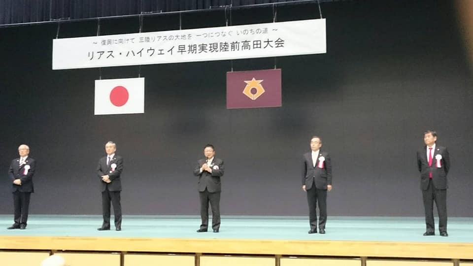 「リアス・ハイウェイ早期実現陸前高田大会」と書かれた横断幕と日本国旗が吊るされた舞台上で、マイクを持った市長を中心に胸章を着けた関係者4名が横一列に並んでいる写真