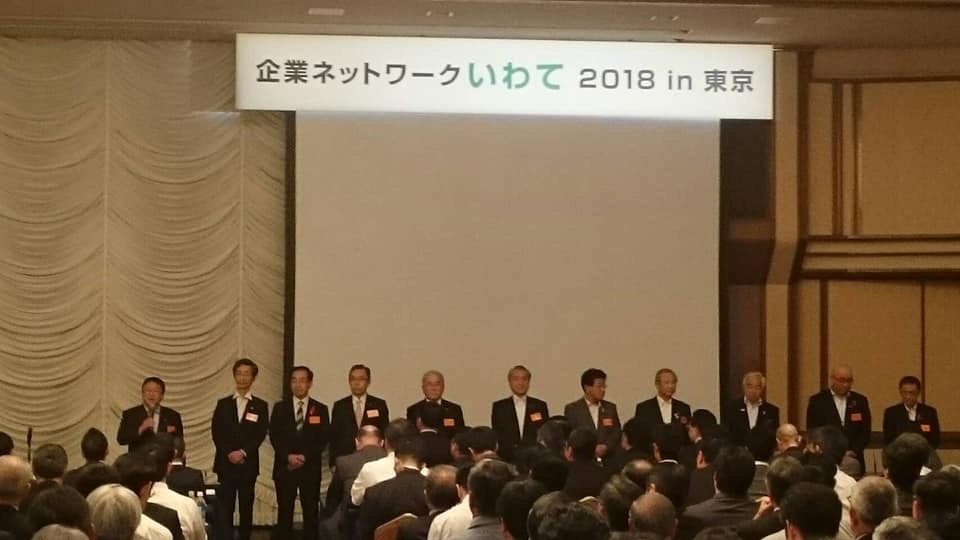 「企業ネットワークいわて 2018in東京」と書かれた横断幕が吊るされた会場で、椅子に座る参加者の前方で横一列に並んで立つ市長と10名の関係者の方々の写真