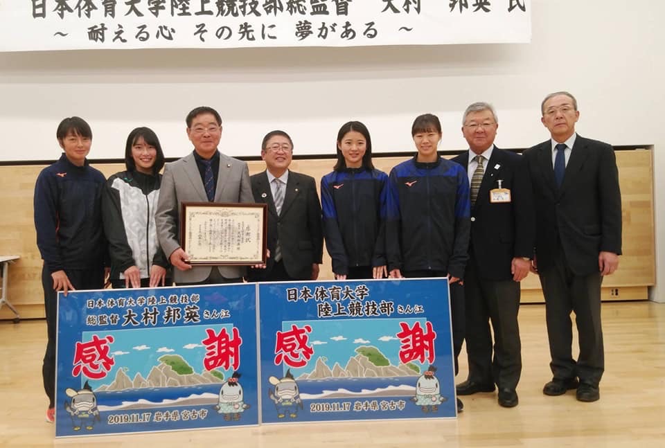 「感謝」という文字と海の絵が書かれている看板を足元に置いて、その後方で横一列に並ぶ市長と日本体育大学陸上競技部の学生や関係者の方々の記念写真