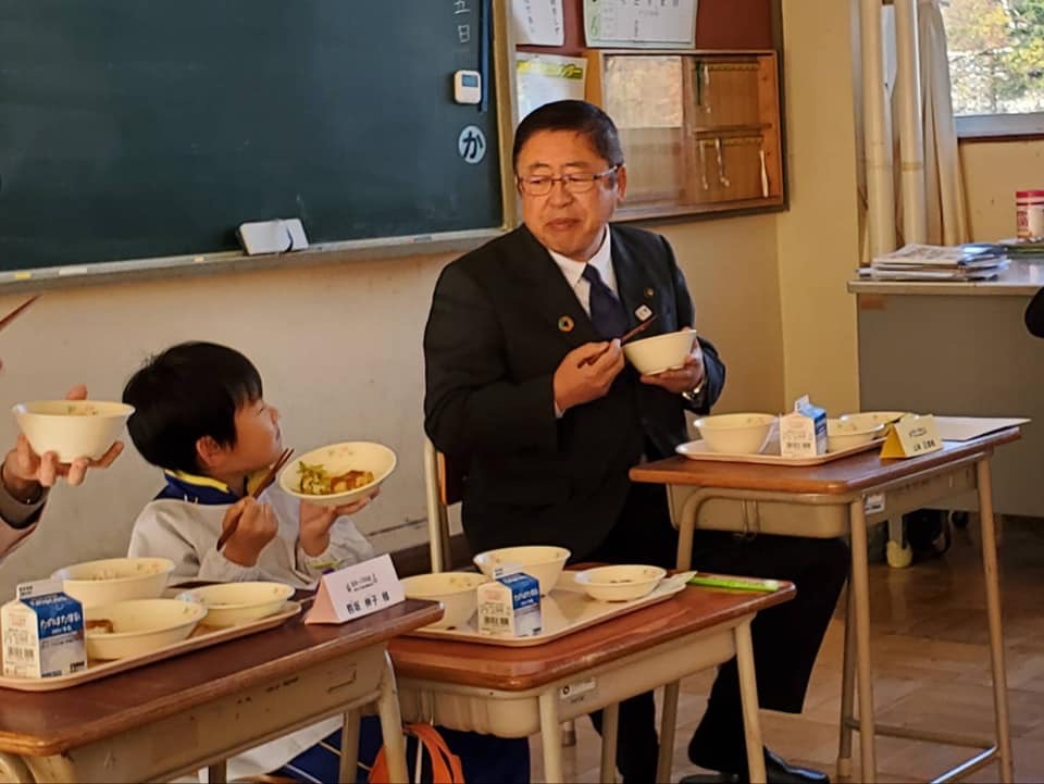 黒板の前に置かれた席に座り給食を食べる様子の市長と男の子の写真