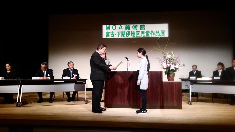 ライトアップされた舞台上で、演台の前に立ち賞状を読み上げている様子の市長と、その向かい側に立つ女の子を写した写真