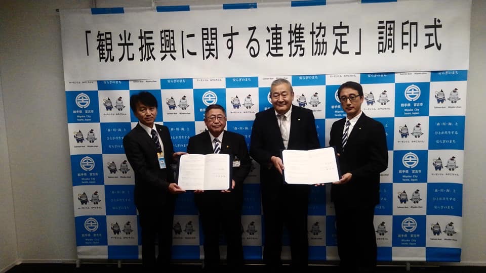 「「観光振興に関する連携協定」調印式」と書かれた横断幕が貼られたバックパネルの前で、協定書をそれぞれ一緒に持つ市長と男性、白髪の男性と眼鏡をかけた男性の記念写真