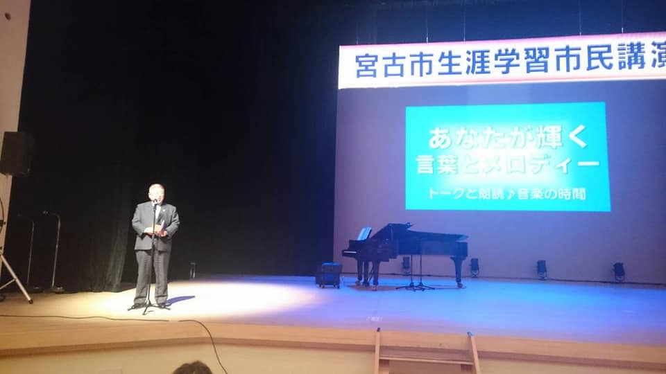 画面が映されたスクリーンとグランドピアノが置かれた舞台の上で、ライトアップされた市長がスタンドマイクに向かい話をしている写真