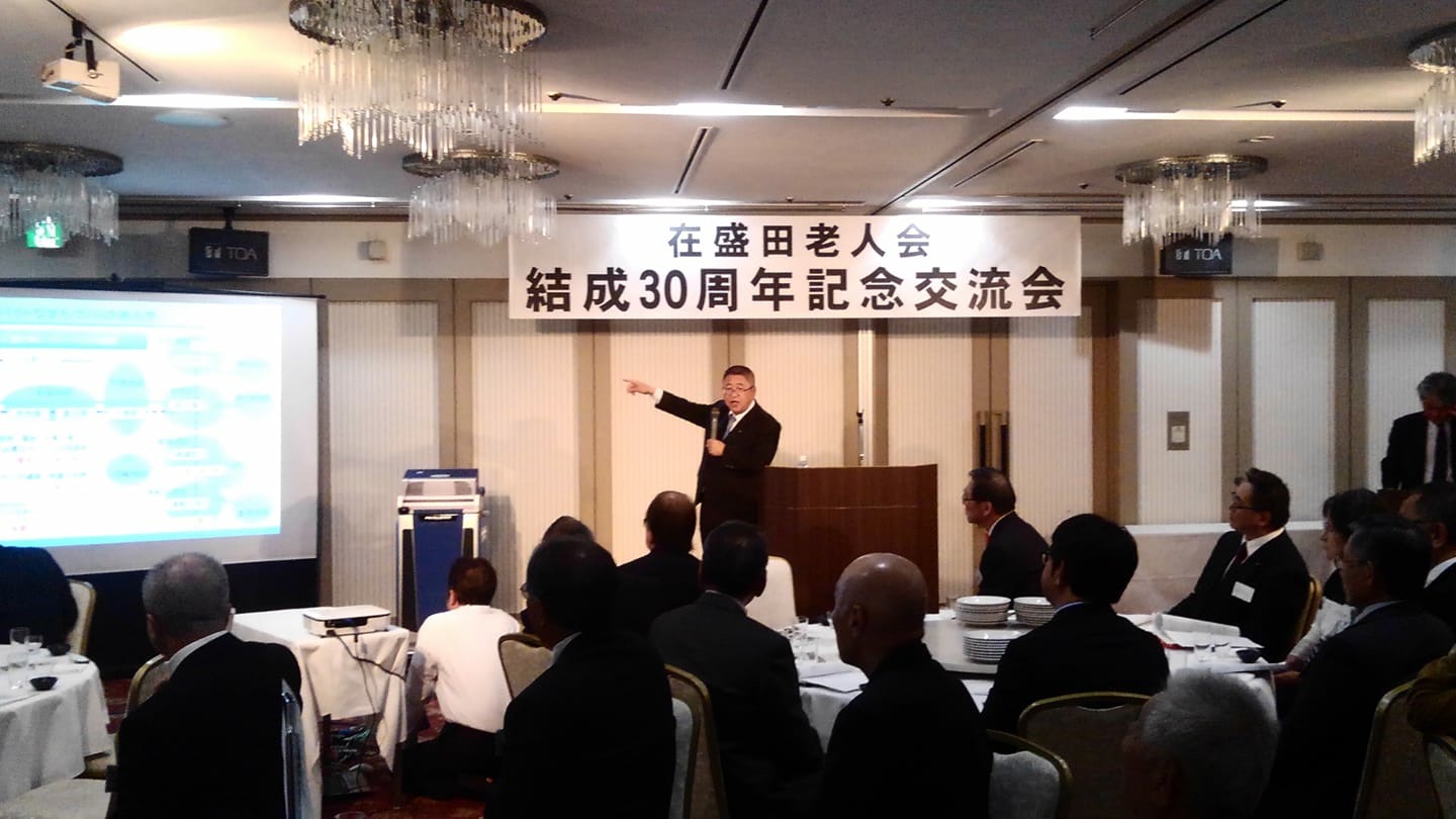 「在盛田老人会 結成30周年記念交流会」と書かれた看板が吊るされた会場で、スクリーンを指さしながらマイクを持って話をする市長と、円卓の席に着きスクリーンを見ながら話を聞く様子の参加者たちの写真
