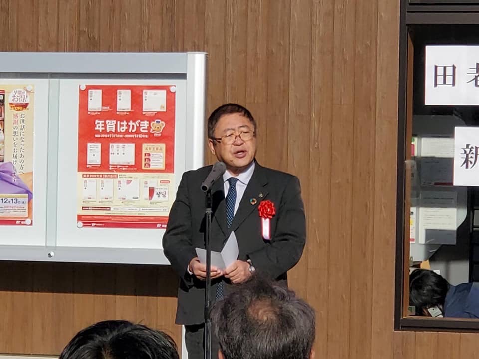 赤い胸章を着けた市長がスタンドマイクに向かって話をする様子の写真