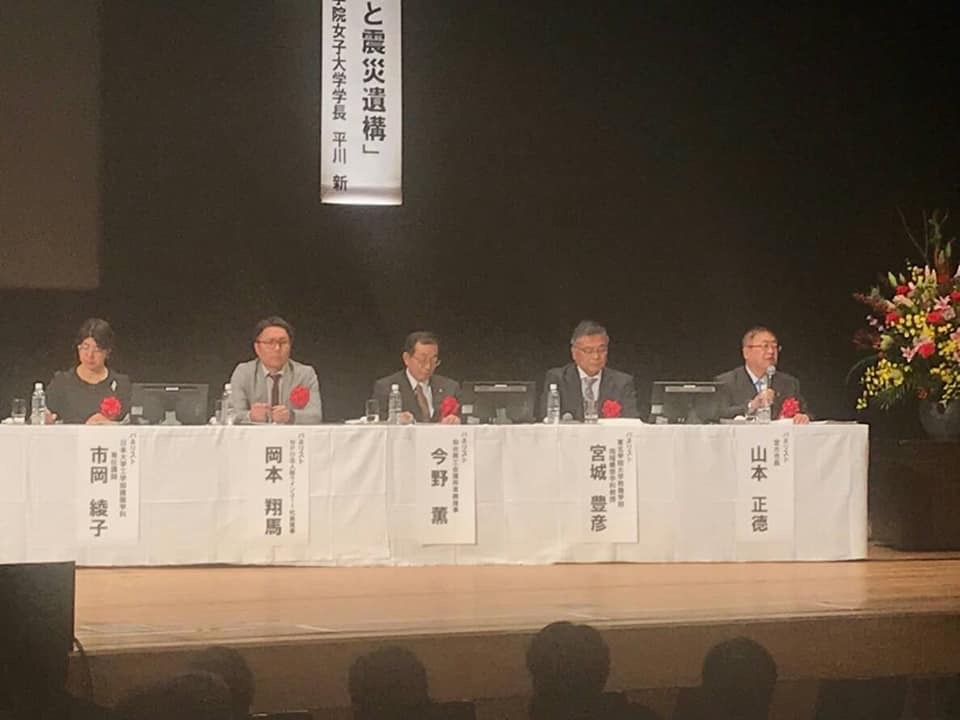 正面に名前札が貼られた白いテーブルクロスが敷かれた長机の席に、赤い胸章をつけた市長と5名の関係者が座っている写真