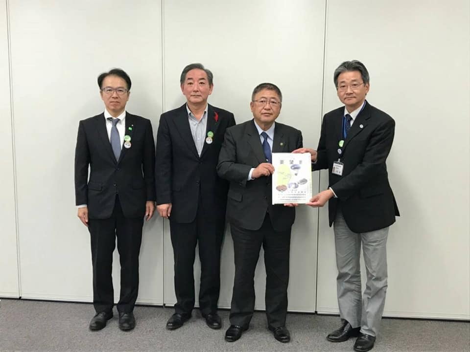 壁の前で要望書を一緒に持つ市長と男性と関係者2名の男性が横一列に並んでいる記念写真