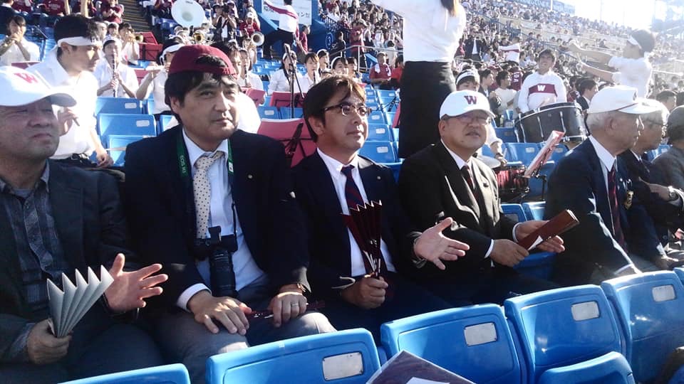野球場のスタンド席に設置された青い椅子に座り、右手に張扇を持ち応援する様子の市長と関係者の方々を写した写真
