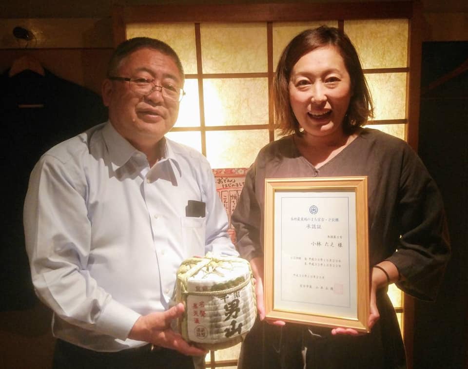 「男山」と書かれた日本酒の小さい樽を持つ市長と、額縁を両手で持ち正面に向ける女性が並んでいる記念写真