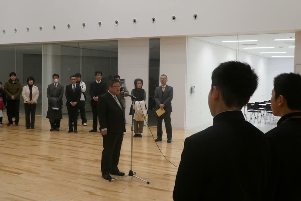 保護者や関係者が見守るなか、中央に設置されたマイクスタンドの前に立つ市長の話を、多良間島へ出発する学生服を着た男子中学生が聞いている後ろ姿の写真