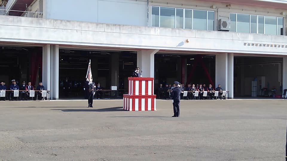 消防署の敷地内で、紅白幕が囲われた台の上に所長が立ち、1名の消防団員が向かい合って立ち敬礼をしている出初式の様子の写真