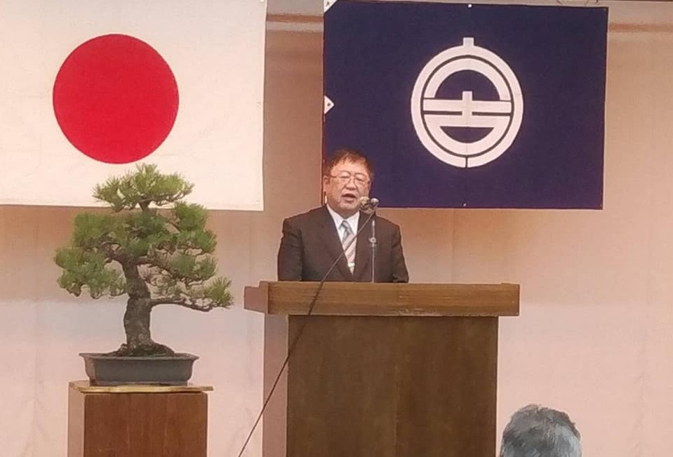 国旗と市旗が掲げられ左側に立派な松の木が置かれた壇上で、市長が挨拶を行っている新年交賀会の様子の写真