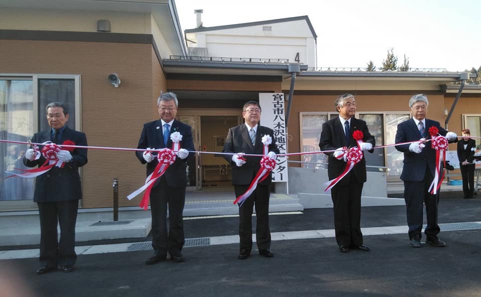 八木沢地区センターの屋外で、市長を中央に4名の関係者が横一列に並びテープカットが行われようとしている開所式の様子の写真