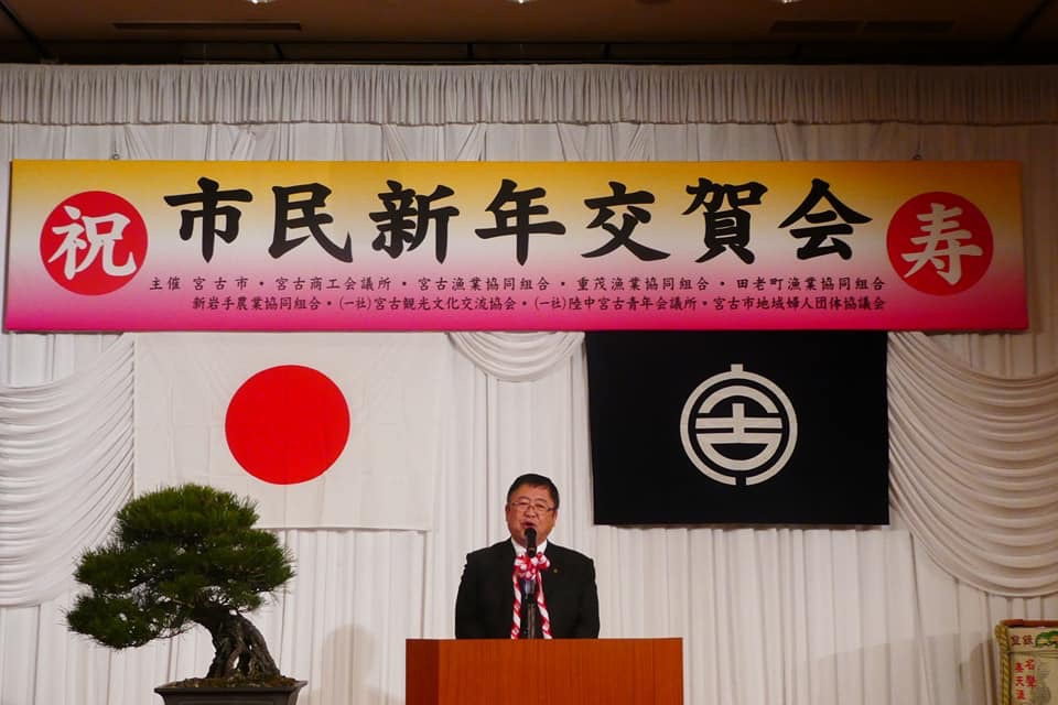 「市民新年交賀会」と書かれた横断幕と国旗と市旗が掲げられ、左側に立派な松の木が設置された壇上で挨拶を行っている市長の写真