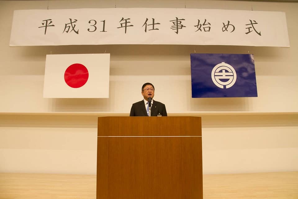 2枚の国旗と市旗「平成31年仕事始め式」と書かれた横断幕が掲げられた檀上で、挨拶を行っている市長の写真