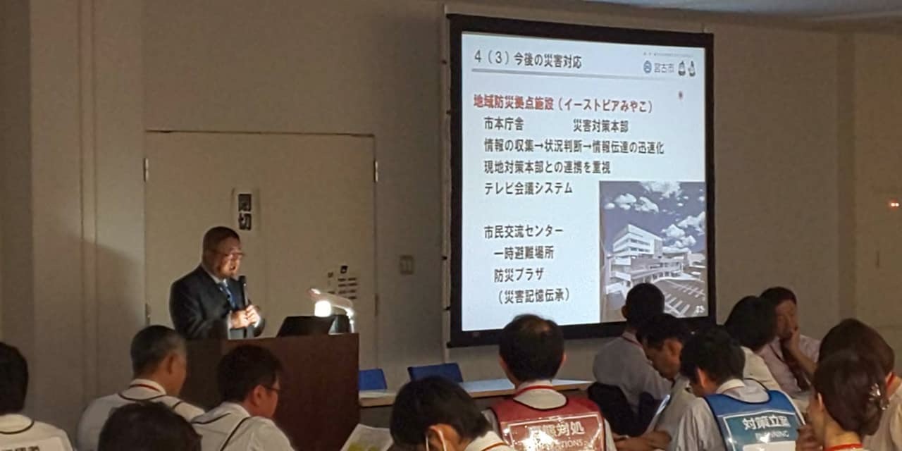 画面が映されたスクリーンの横の演台で、両手でマイクを持ち話をする様子の市長の写真