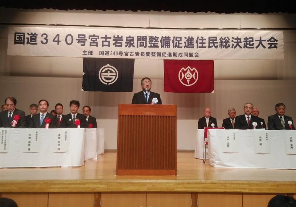 「国道340号 宮古岩泉間整備促進住民総決起大会」と書かれた横断幕が書かれた舞台上で、名前札が掛けられたテーブルの席につく関係者たちの間に置かれた演台で話をする様子の市長の写真