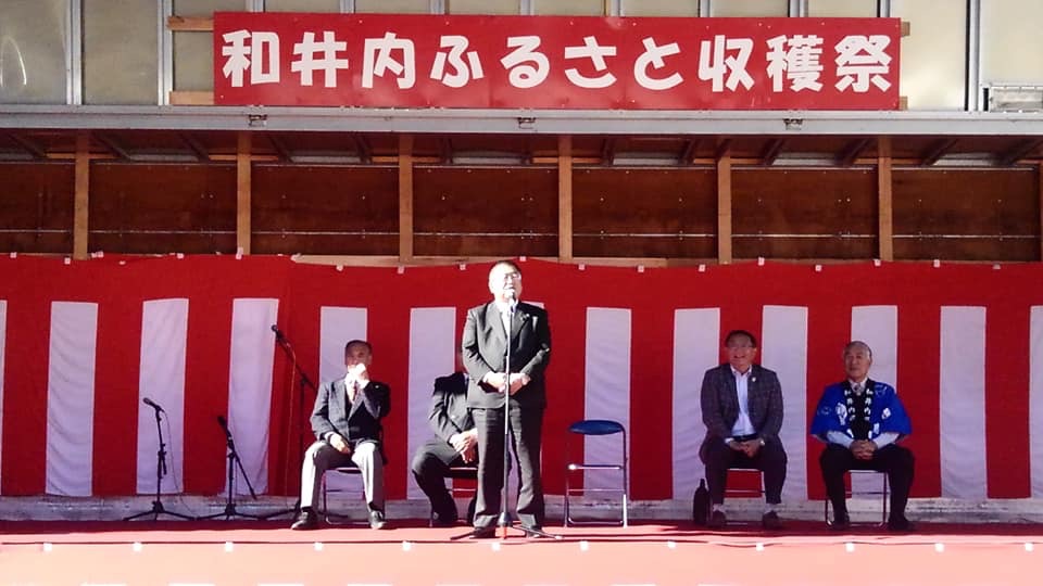 「和井内ふるさと収穫祭」と書かれた赤い看板の下で、スタンドマイクに向かい話をする様子の市長の写真