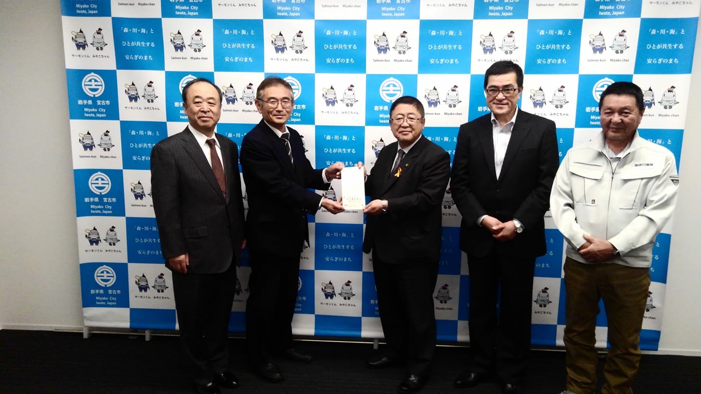 バックパネルの前で寄附金の封筒を持つ市長と男性を中心に3名の男性が横一列に並んでいる記念写真