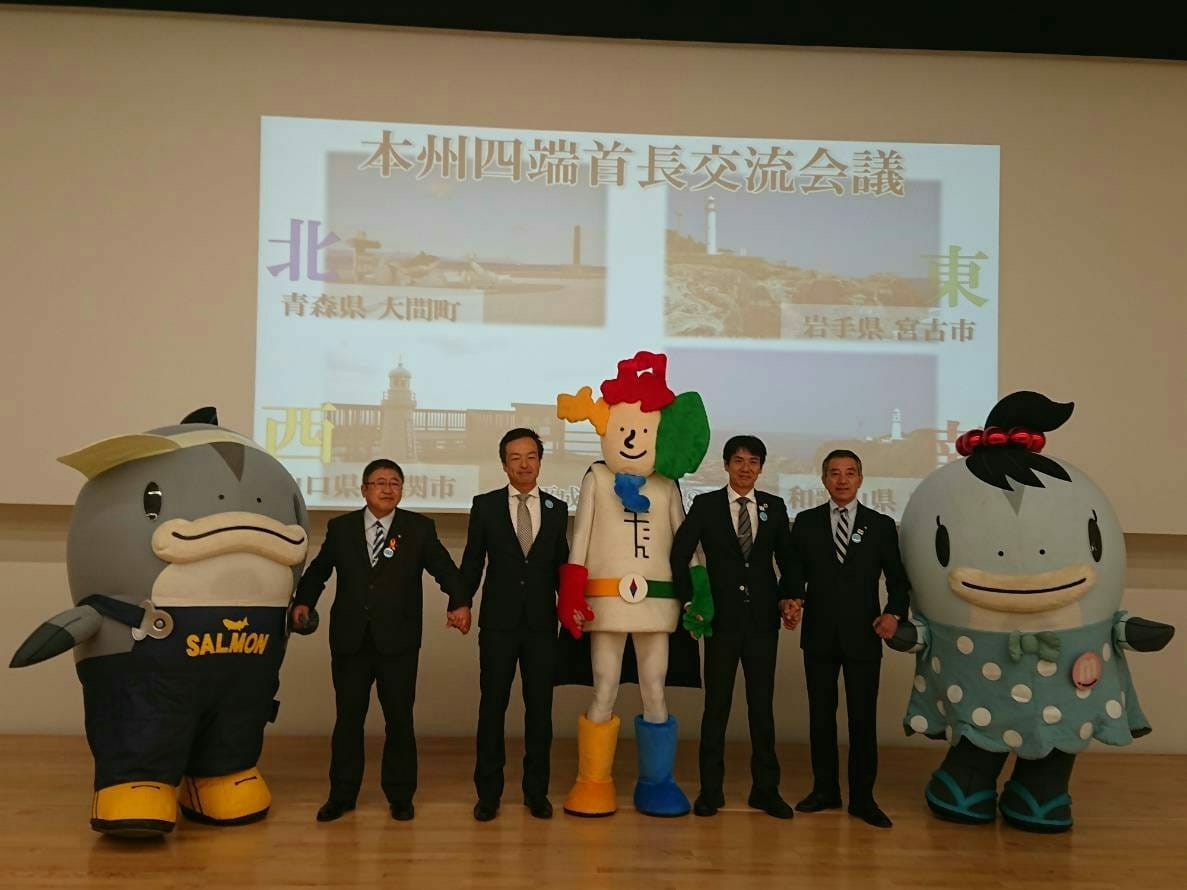 画像が映し出されたスクリーンの前で横一列に並んで記念撮影をする山本市長、山口県下関市の前田晋太郎市長、和歌山県串本町の田嶋勝正町長、青森県大間町の金澤満春町長、サーモンくんとみやこちゃん、よんたんのの写真