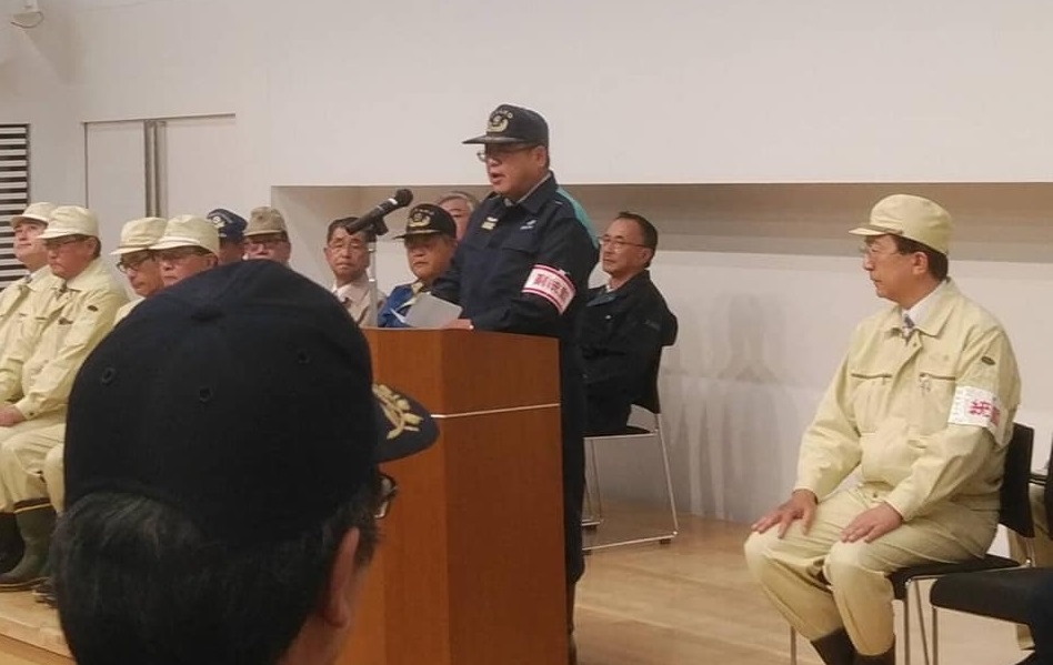 薄い黄色の作業服と帽子を身に着けた関係者たちの中心に置かれた演台で話をする様子の市長の写真
