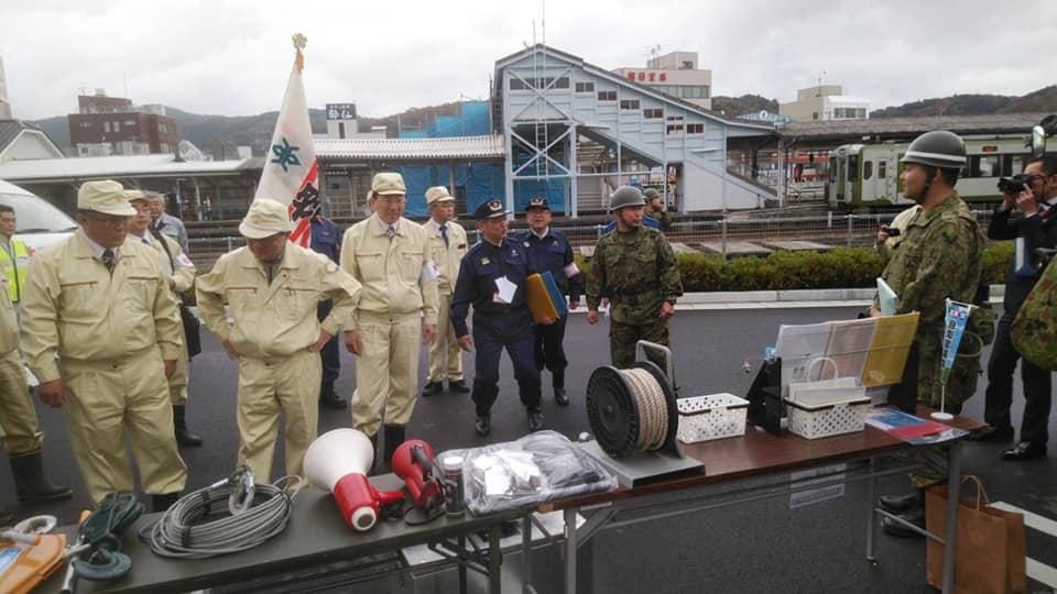 コードや拡声器などが置かれた長机の後方で、薄い黄色の作業服と帽子を身に着けた関係者や迷彩服を着た自衛隊員たちが立っている様子の写真