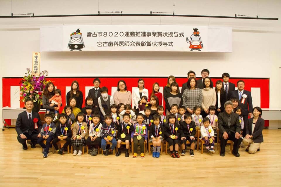 「宮古市8020運動推進事業賞状授与式 宮古歯科医師会表彰賞状授与式」と書かれた横断幕が吊るされ、壁に紅白幕が貼られた会場で、椅子に座る子供たちや後方に立つ保護者の方々が並んで写っている記念写真