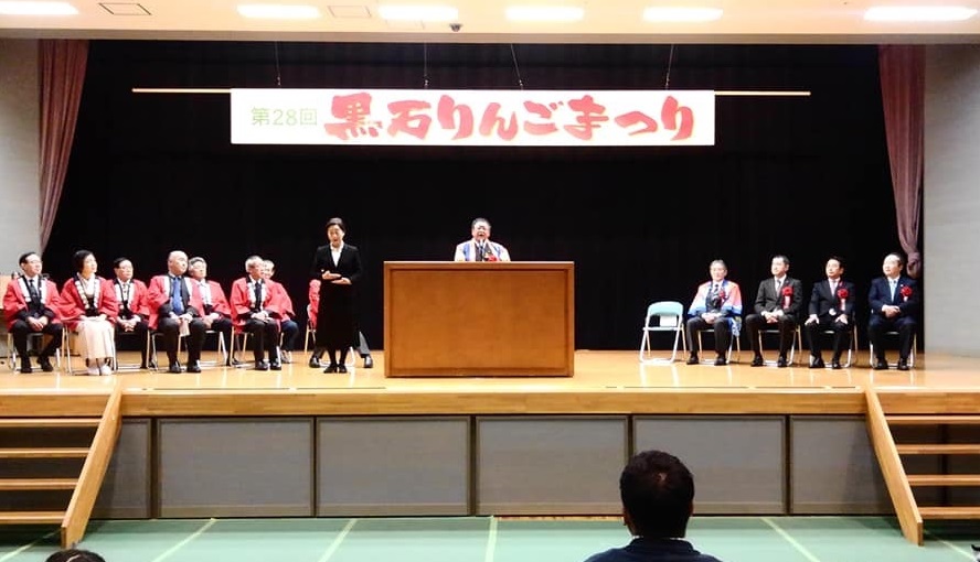 「第28回黒石りんごまつり」と書かれた横断幕が書かれた舞台の中心に置かれた演台で話す様子の市長と、演台の両側に並べられた椅子に座る関係者の方々の写真