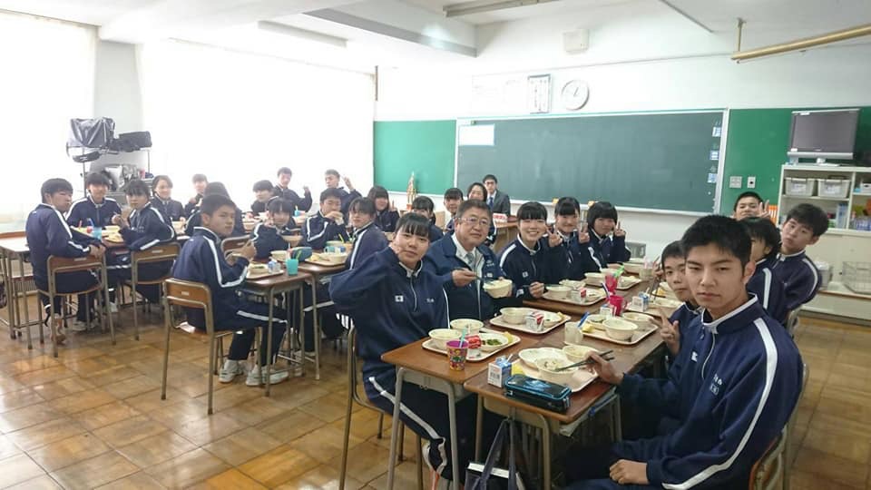 教室で机を向かい合わせにして給食を食べる様子のジャージを着た中学生たちと市長の写真