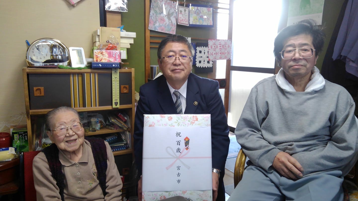 熨斗のついた祝い品の箱を持った市長を中心に、満100歳を迎えた小笠原ヨシさんとご家族の男性が写っている記念写真