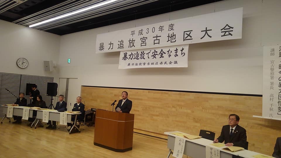 「平成30年度 暴力追放宮古地区大会」と書かれた横断幕が掲げられた会場の中央で、演台に置かれたマイクに向かい話をする様子の市長と、演台の両側に並べられたテーブルの席につく関係者たちの写真