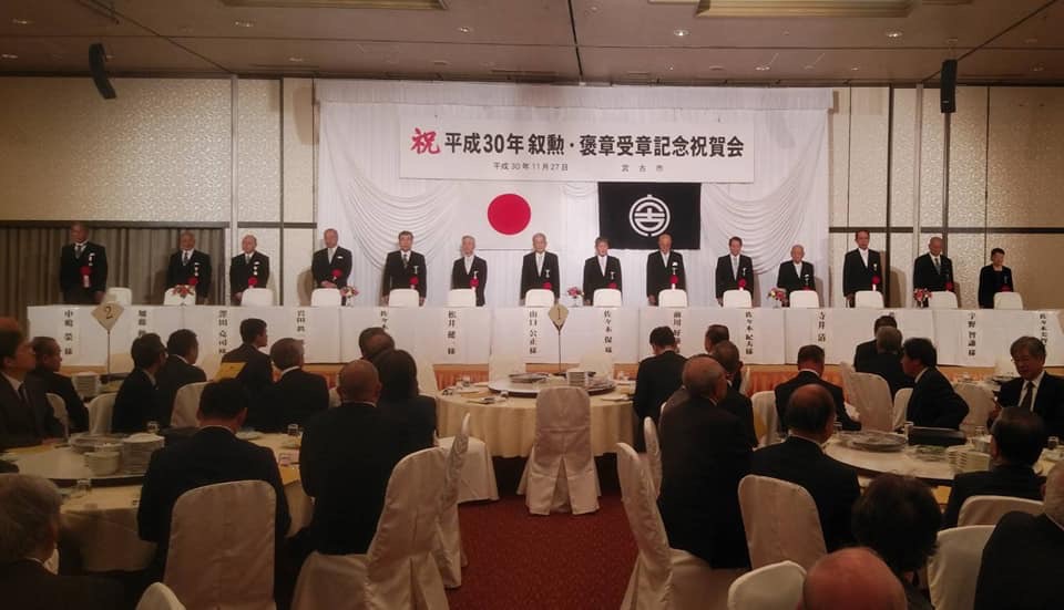 「祝 平成30年勲章・褒章受章記念祝賀会」と書かれた横断幕と日本国旗、宮古市の市町村旗が掲げられた舞台上に置かれたクロスが敷かれたテーブルと椅子の後方に立つ14名の関係者の方たちと、舞台の下に並べられた円卓の席に着く関係者の方々の写真