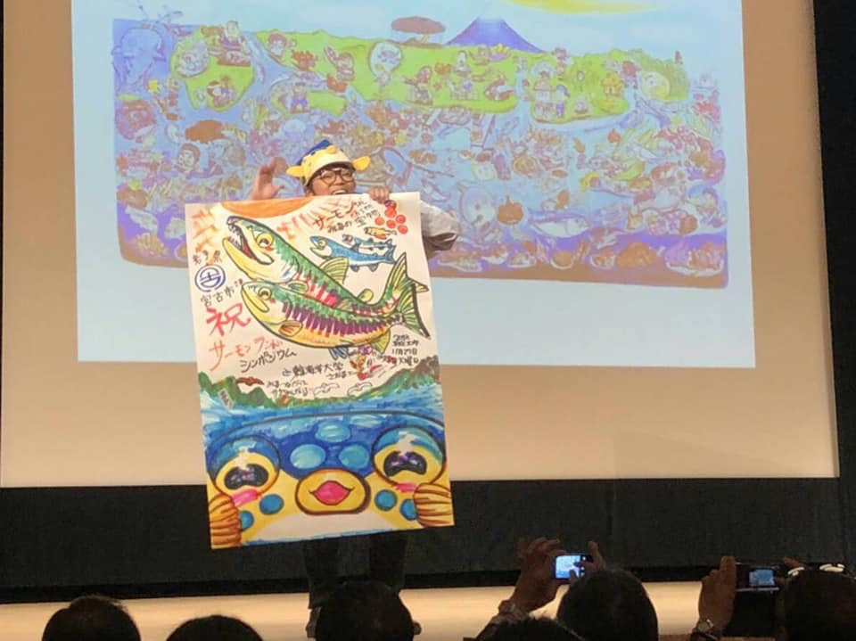 スクリーンの前でサーモンのカラフルな絵を描いたポスターを両手で持つ様子のさかなクンの写真
