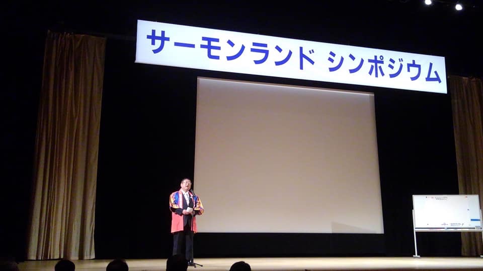 「サーモンランドシンポジウム」と書かれた横断幕が掲げられた舞台上で、法被を着てスタンドマイクに向かい話をする様子の市長の写真