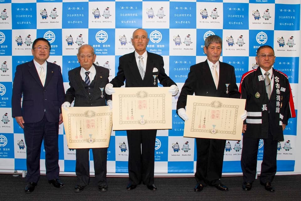 バックパネルの前で症状を持つ3名の男性と、両側に立つ市長、法被を着た男性が横一列に並んでいる記念写真