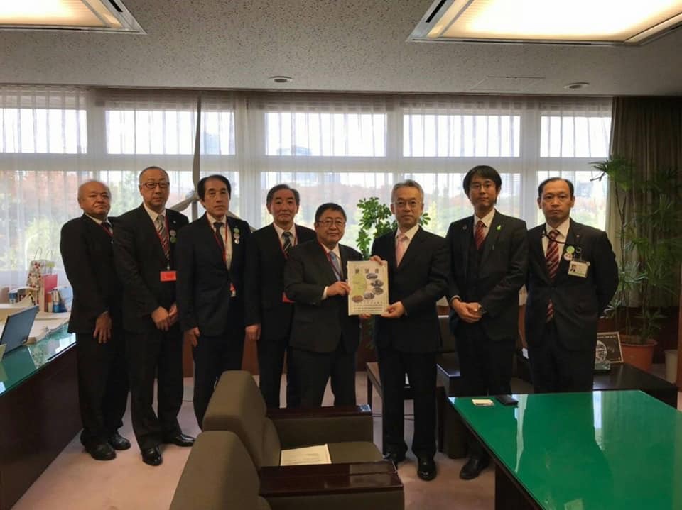 右側の緑色の机の後ろで要望書を一緒に持つ市長と男性を中心に、関係者の男性6名が横一列に並んでいる記念写真