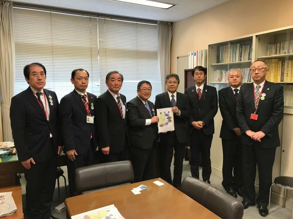 茶色の机の後ろで要望書を一緒に持つ市長と男性を中心に、関係者の男性6名が横一列に並んでいる記念写真