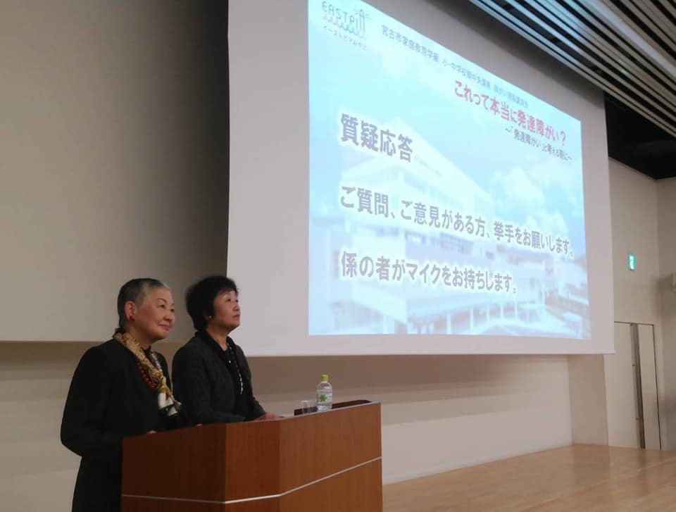 質疑応答の画面が映されたスクリーンの横に置かれた演台に立つ2名の女性を写した写真