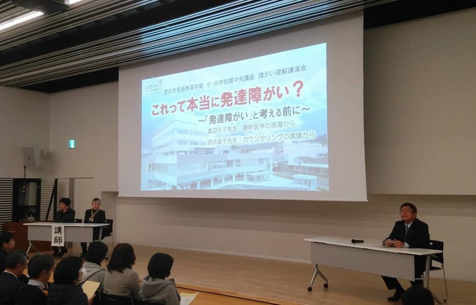 「これって本当に発達障がい？」という文字の背景に建物が写った画面が映されたスクリーンの横に置かれた席に座る市長とスクリーンを見ている様子の参加者たちの写真