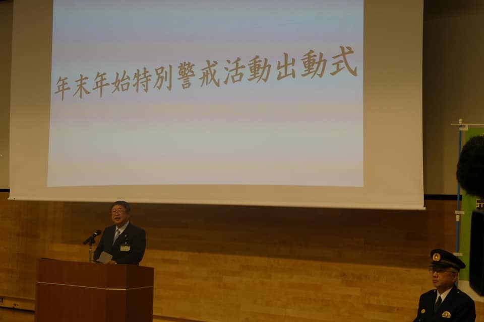 「年末年始特別警戒活動出動式」という文字が映されたスクリーンの前の演台に置かれたマイクに向かい話をする様子の市長の写真と、スクリーンの横の椅子に座る制帽と制服を身に着けた警察官の写真