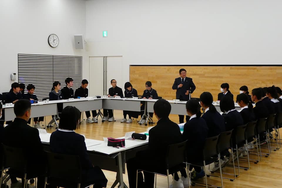 ロの字型に並べられたテーブルの席に着く制服を着た男女の中学生たちと、一番前で立ってマイクを持ち話をする様子の市長の写真