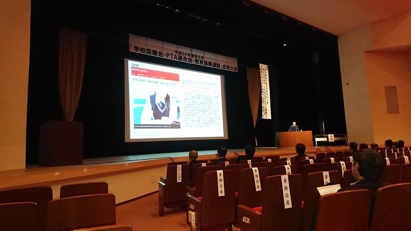 横断幕とスクリーンが吊るされた舞台を見る様子の椅子に座った参加者たちの写真