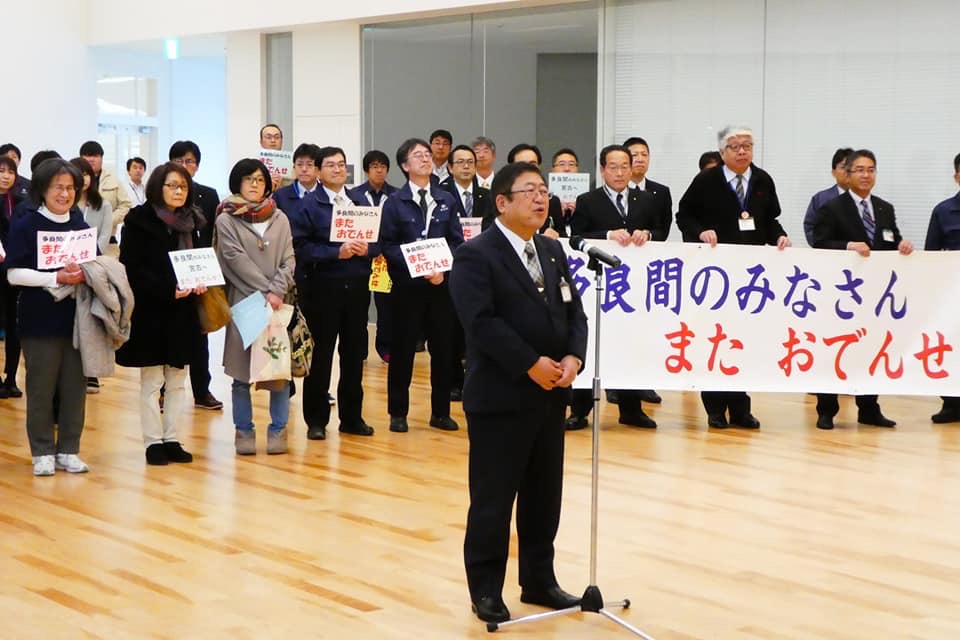 「多良間のみなさん またおでんせ」と書かれた横断幕やプラカード持って並ぶ参加者の方々の前でスタンドマイクに向かい話をする様子の市長の写真