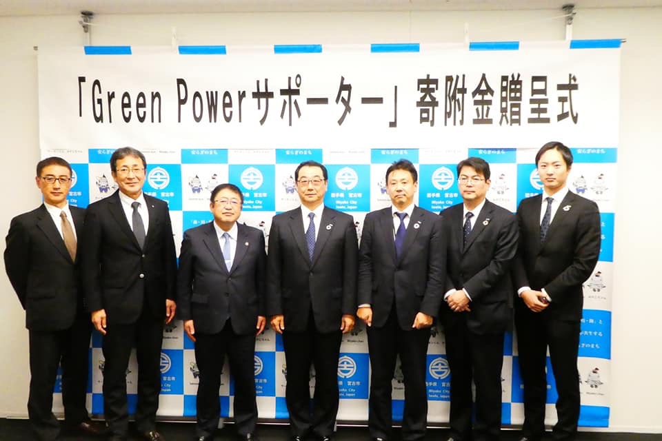 「「Green Powerサポーター」寄附金贈呈式」と書かれた横断幕が貼られたバックパネルの前で横一列に立って並ぶ市長と6名の男性の写真