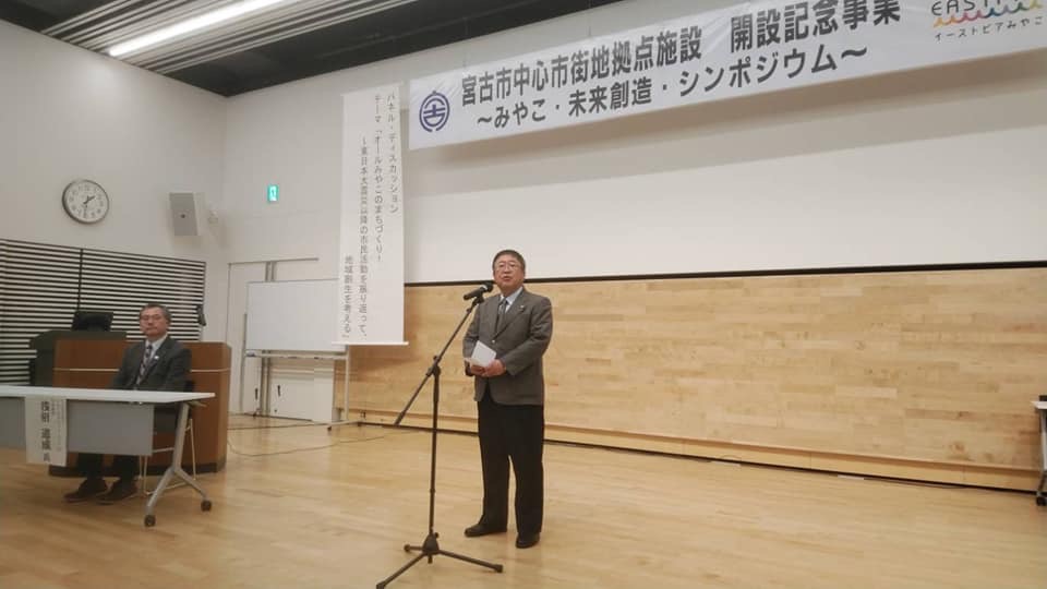 「宮古市中心市街地拠点施設 開設記念事業～みやこ・未来創造・シンポジウム～」と書かれた横断幕が掲げられた会場で、スタンドマイクに向かい話をする様子の市長