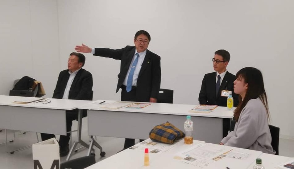 横に並べられた長机に座る2名の男性の間に立ち右手を広げて参加者の女性に説明をする様子の市長の写真