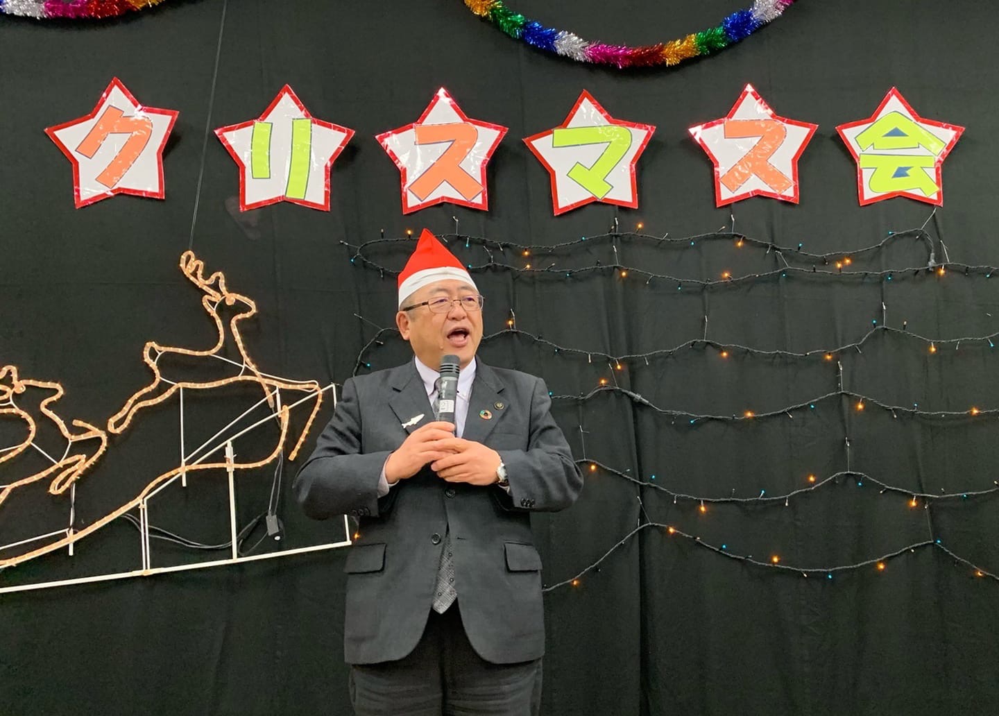 クリスマスの装飾が施された黒い壁の前で、サンタの帽子を被り両手にマイクを持って話をしている様子の市長の写真