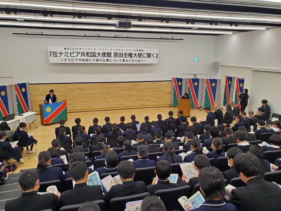 「在ナミビア共和国大使館 原田全権大使に聞く」と書かれた横断幕が吊るされた会場で、演台の前に立つ原田大使と資料を見ながら話を聞く様子の学生たちの写真