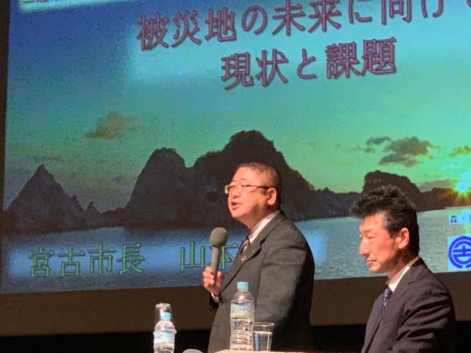 画面が映されたスクリーンの前でマイクを持ち話をする様子の市長と、その隣で椅子に座っている男性の写真